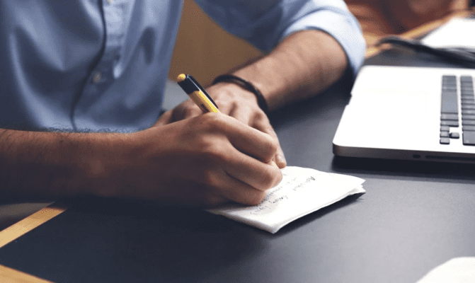 Person writing in a notepad in front of a laptop on a desk