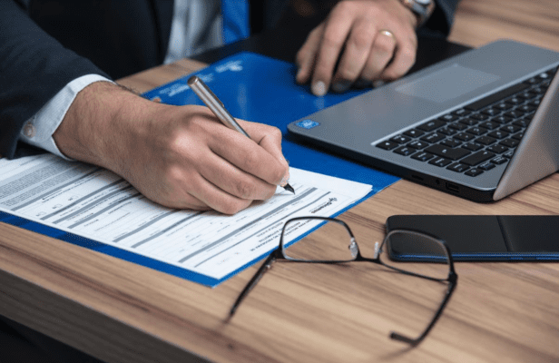 Photo of a man's hands signing a contract. He wears a suit. A laptop, cell phone and pair of glasses sit on a wooden desk.